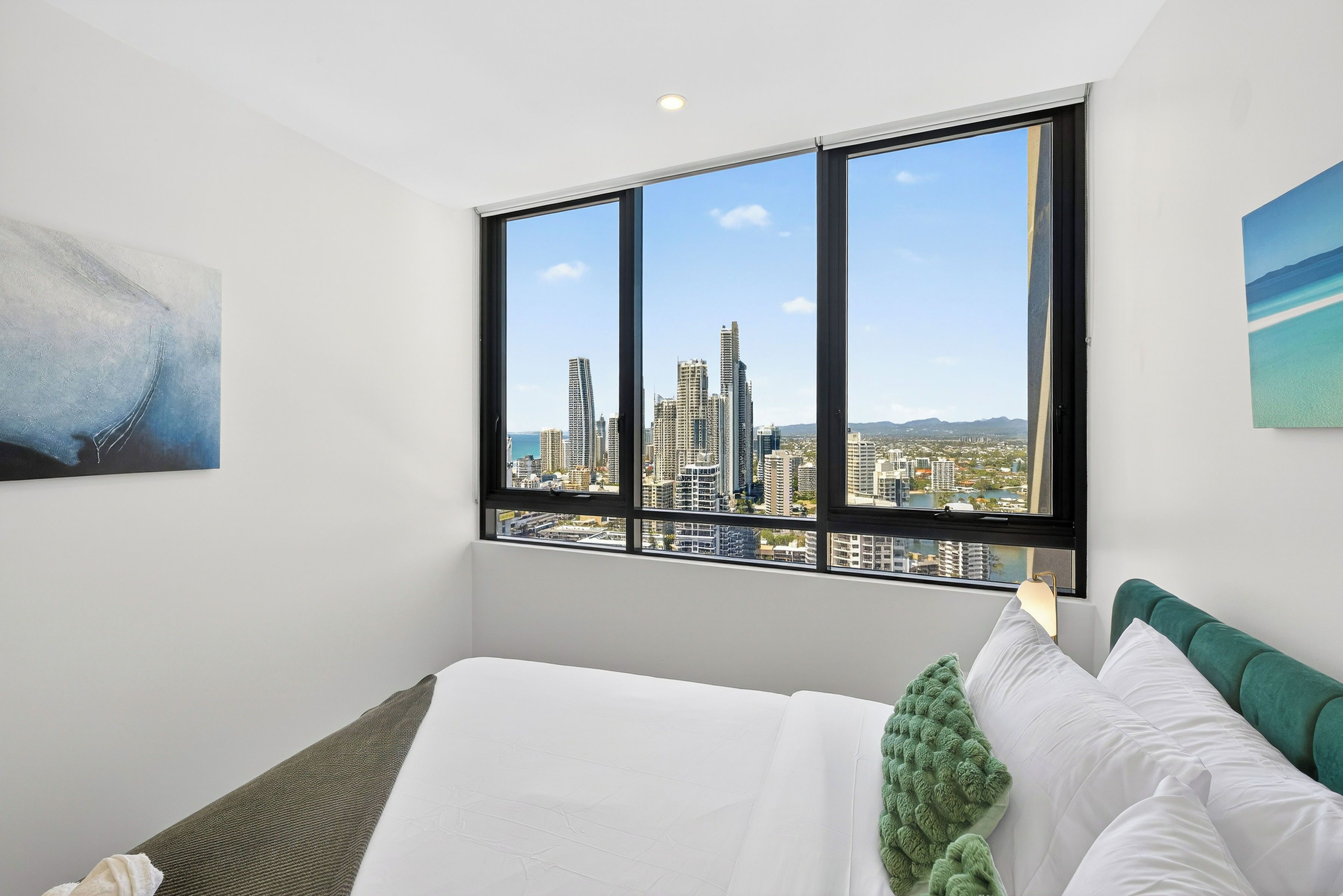 A hotel room with a large window offering a cityscape view, a neatly made bed, and a painting on the wall.