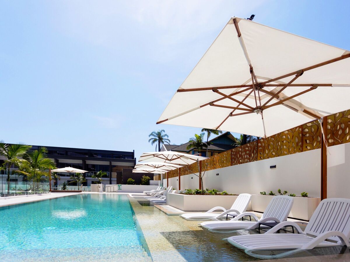 A swimming pool with lounge chairs and umbrellas, surrounded by a modern building with palm trees.