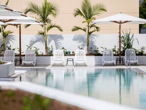 Un espace piscine avec des chaises et des parasols blancs, entouré de palmiers et d’un mur de couleur beige.