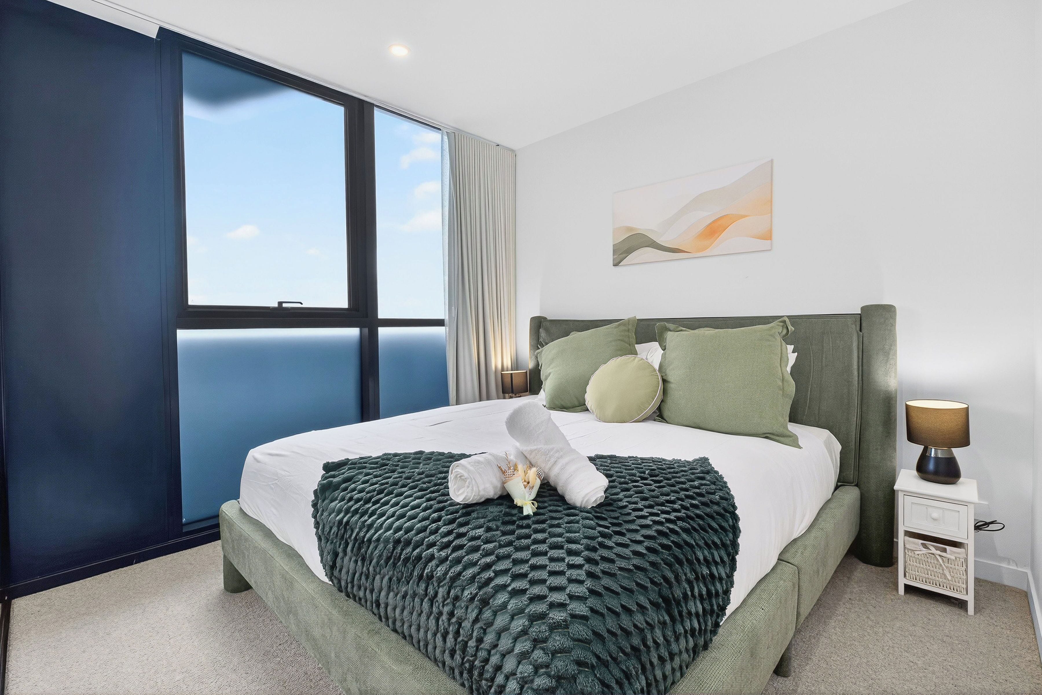 A modern hotel room with a large bed, a nightstand, a wall-mounted lamp, and a window with a view of the sky.