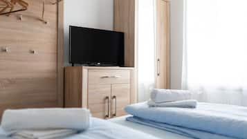 Ein Hotelzimmer mit einem Holzschrank, einem Flachbildfernseher darauf, einem Bett mit blauer Bettwäsche und einem Fenster mit durchsichtigen Vorhängen.