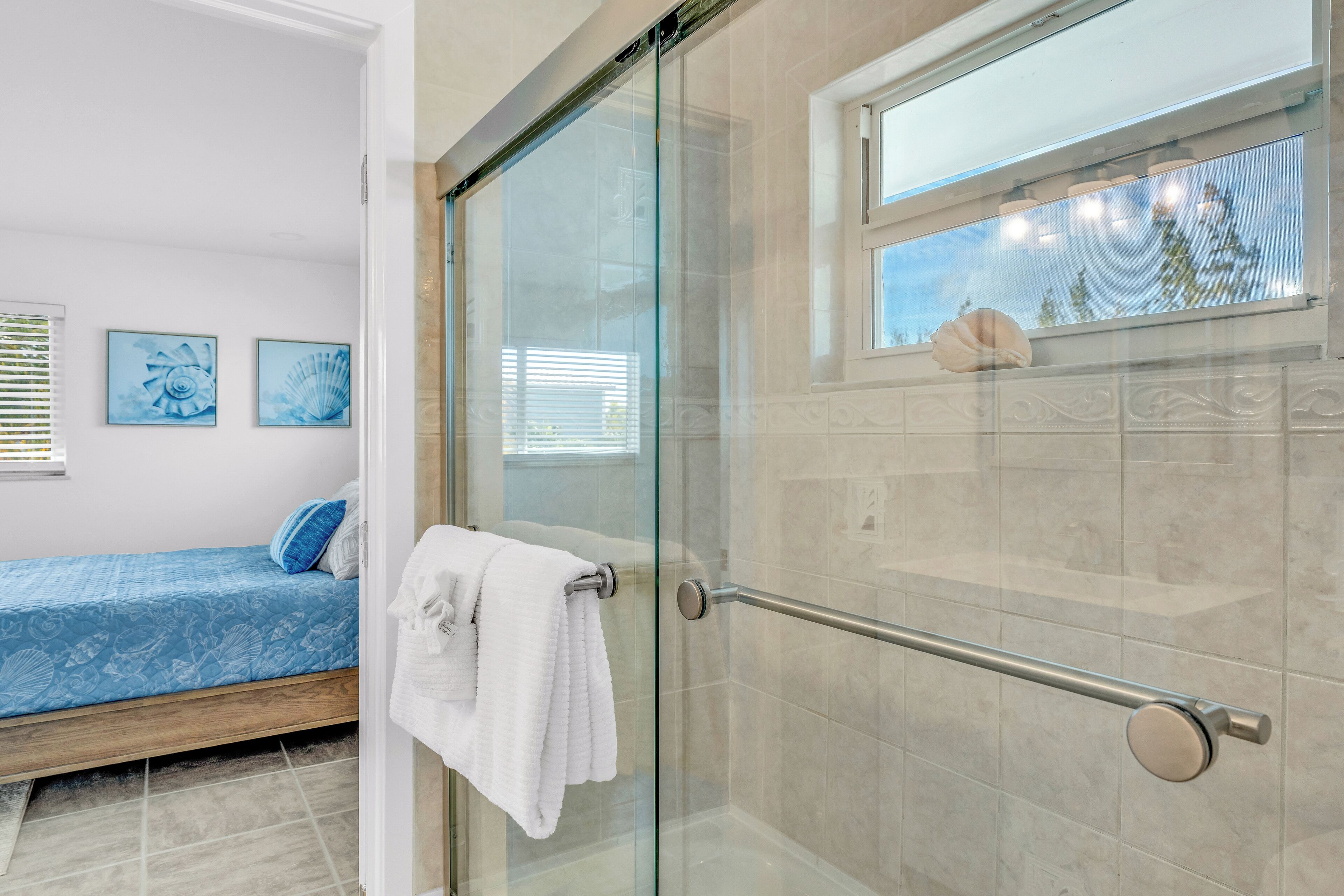 A modern bathroom with a glass shower enclosure, a bed with blue bedding, and artwork on the wall.
