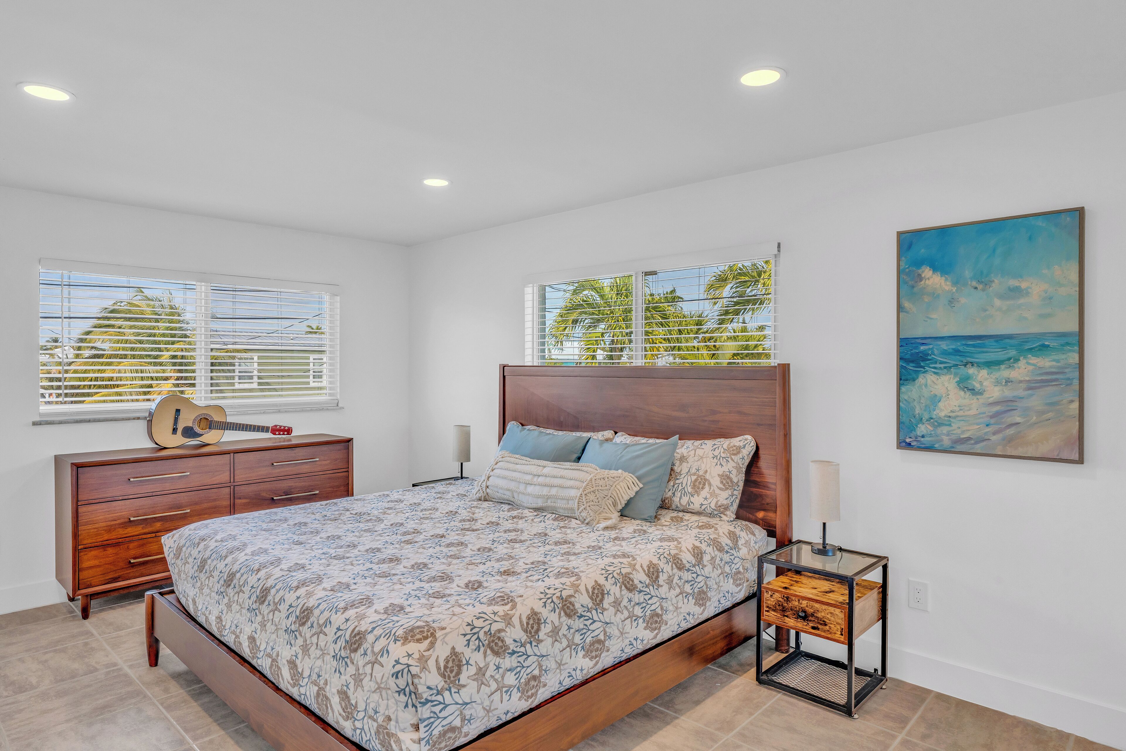 A bedroom with a wooden bed, a nightstand, a guitar, and a painting of the ocean.
