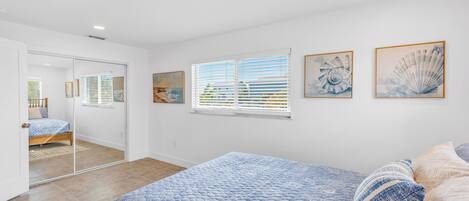 Une chambre avec un grand lit, une fenêtre avec des stores et deux œuvres d’art encadrées accrochées au mur.