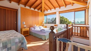 Une chambre avec un grand lit, un plus petit lit, des poutres en bois au plafond, un balcon avec une balustrade et une vue sur les arbres.