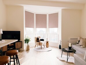 Ein modernes Wohnzimmer mit Fernseher, Sitzgelegenheiten, einer Pflanze und Blick durch große Fenster.