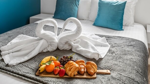 Un lit bien fait, avec des oreillers blancs et bleus, une couverture grise et un plateau de petit-déjeuner contenant des croissants, des fruits et des raisins.