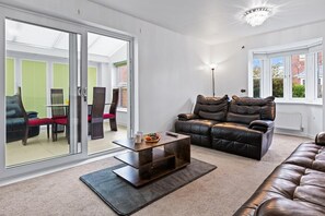 A modern living room with a leather sofa, a wooden coffee table, and a glass door leading to an outdoor seating area. - Spacious Comfortable 4B Home w en-suites (Great Sankey)