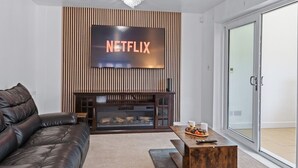 Un salon moderne avec un canapé en cuir, une table basse en bois et une cheminée. Un grand téléviseur fixé au mur affiche le logo de Netflix.