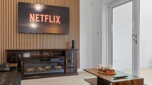 Un salon moderne avec une cheminée, un écran affichant le logo de Netflix, un canapé en cuir et une table basse sur laquelle sont disposés des en-cas.