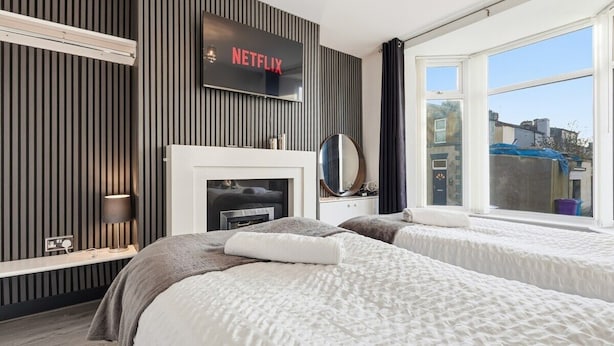 A modern hotel room with two beds, a fireplace, a TV mounted on the wall, and a large window offering a view of the city. - The Crews Quarters Anfield - Sleeps 10 - Modern (Merseyside)