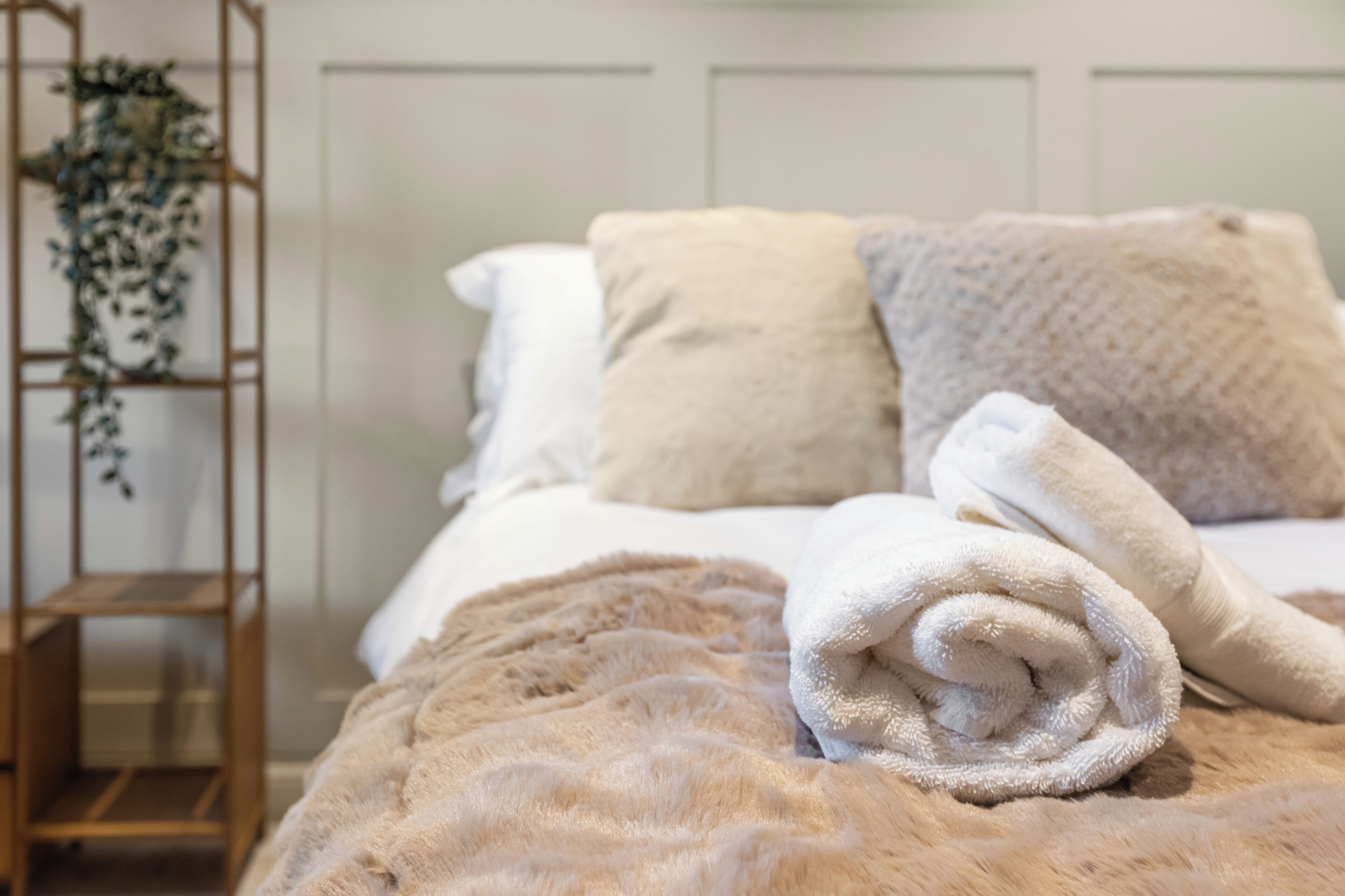 A neatly made bed with a fluffy beige blanket, a rolled white towel, and a wooden shelf with a plant.