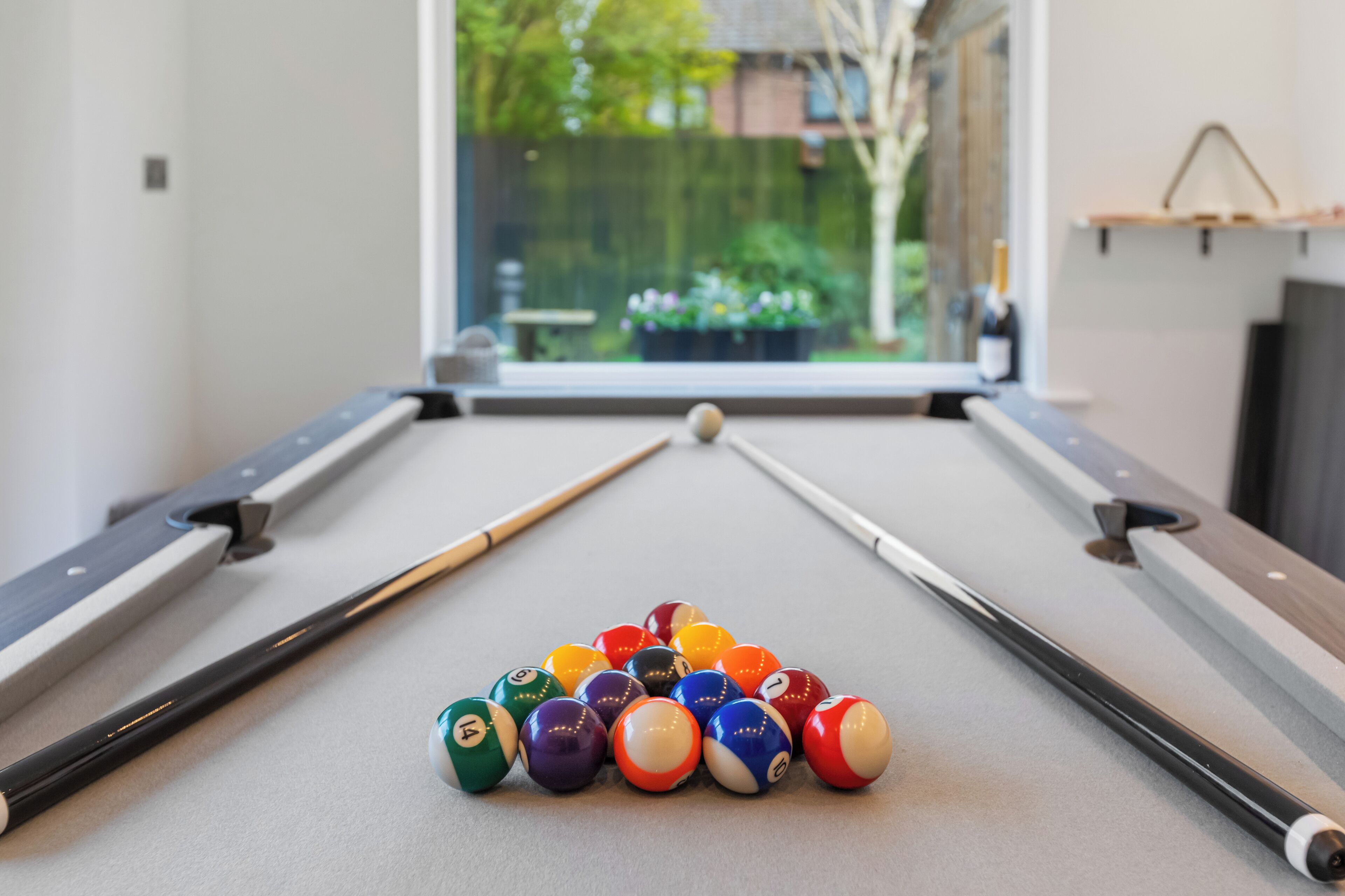 Una mesa de billar con un juego de bolas dispuestas en forma de pirámide, una cola de billar y una ventana con vistas a la vegetación.