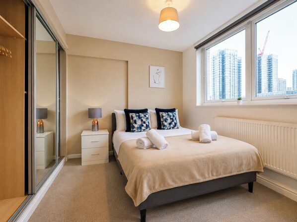 A modern hotel room with a bed, bedside tables, a window with a city view, and a radiator. - Stylish 2 Bed Flat Near Old Street (Greater London)