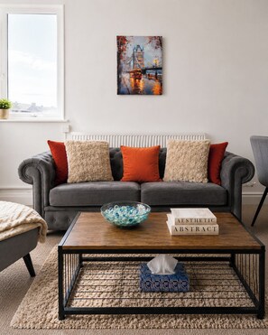 A modern living room with a grey sofa, decorative pillows, a coffee table, and a framed painting of a bridge. - Stylish 2 Bed Flat Near Old Street (Greater London)