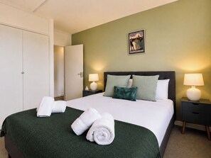 A neatly made bed with white linens and rolled towels, a dark headboard, two bedside lamps, and a framed picture on the wall. - Stylish 2 Bed Flat Near Old Street (Greater London)