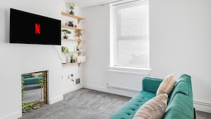 A living room with a teal sofa, a mounted TV displaying the Netflix logo, a window with blinds, and a plant shelf.