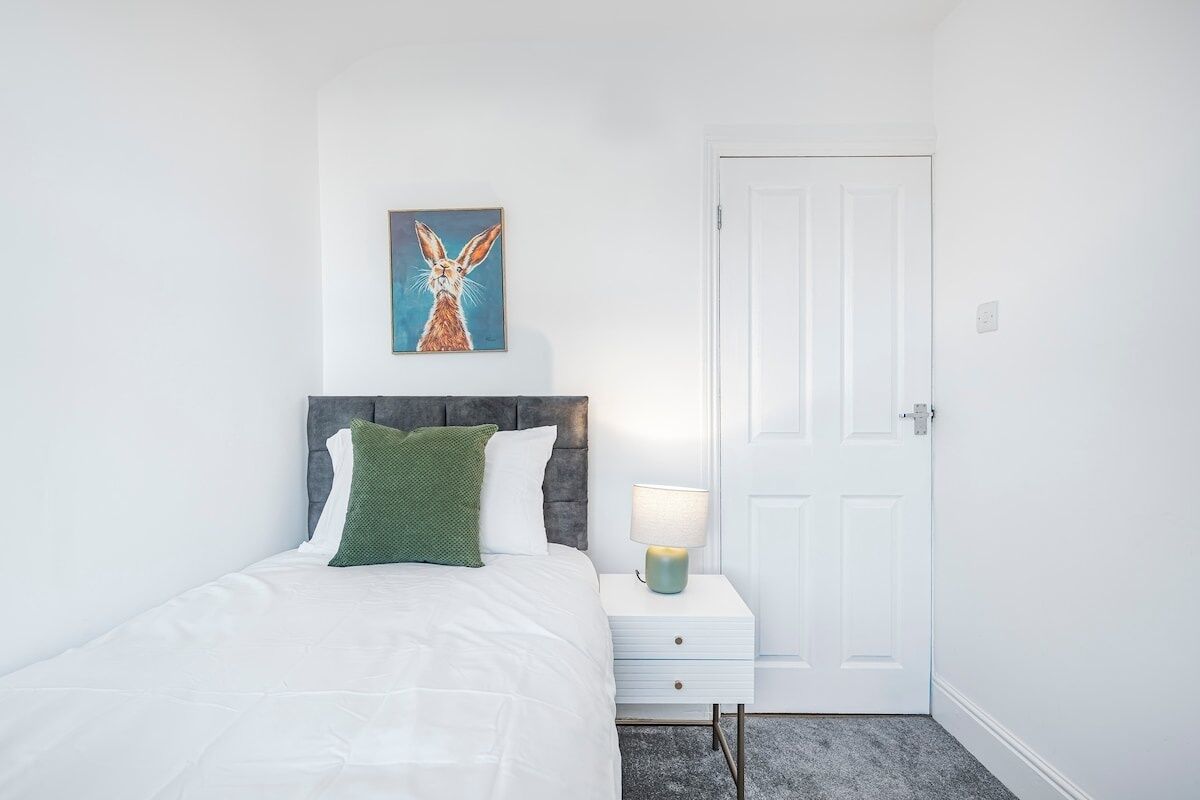 Un dormitorio con cama, mesita de noche, lámpara, puerta y un cuadro en la pared.