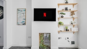 A modern living room with a flat-screen TV mounted on the wall, a dartboard, a framed abstract painting, and a small indoor garden.