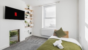 A bedroom with a bed, a TV mounted on the wall, a window with blinds, and a shelf with plants.