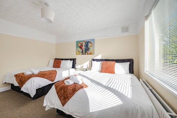 Ein Hotelzimmer mit zwei Betten, beige Wänden, einer Deckenleuchte, einem Elefantenbild und einem Fenster mit Jalousien.