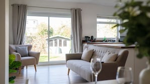 A spacious living room with a sofa and armchair, a dining table with wine glasses, and a kitchen area with a sink and window.