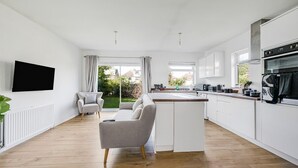 A modern kitchen with white cabinetry, a wooden countertop, and a built-in oven. There is a grey sofa and a dining table with chairs in the adjacent living area.