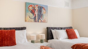 A hotel room with two beds, each with white bedding and orange accents, a painting of an elephant on the wall, and a nightstand with two lamps.