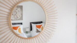 Un miroir rond, doté d’un cadre en forme de soleil, reflète un lit avec une literie blanche et des oreillers orange et bruns, une tête de lit grise et une œuvre d’art accrochée au mur.