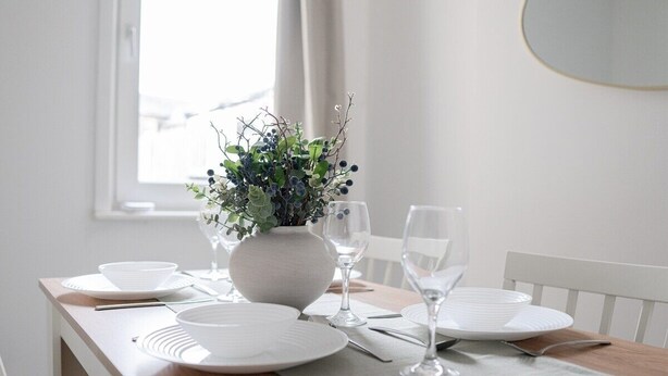 Une table à manger dressée avec de l’argenterie blanche, des couverts et un vase contenant des feuilles vertes.