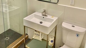 A modern bathroom with a glass-enclosed shower, a white sink with a gold-colored base, and a toilet with a blue sticker.