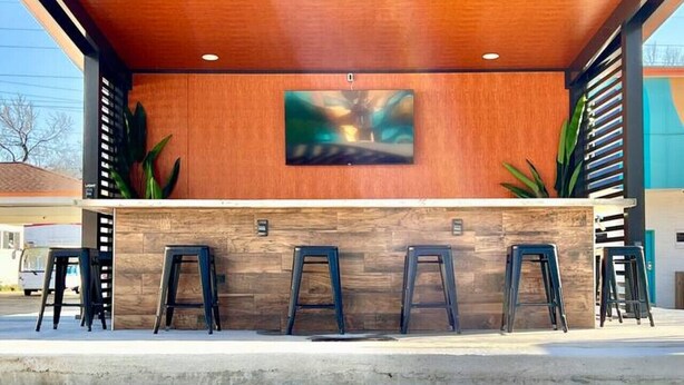 A modern bar area with a wooden ceiling, a flat-screen TV mounted on the wall, and three stools.