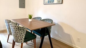 Et spiseområde med et rektangulært bord, seks stole med geometriske mønstre, en potteplante og et billede på væggen.