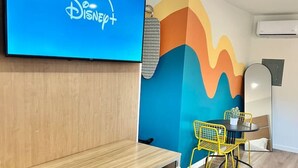 Una habitación con un escritorio de madera, una silla, un televisor que muestra el logotipo de Disney+ y una pared colorida, además de una pequeña mesa con sillas.