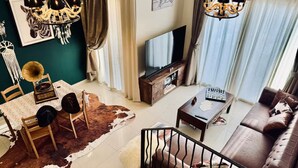 A modern living room with a brown leather sofa, a wooden coffee table, a TV on a wooden stand, a chandelier, and a cowhide rug.