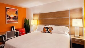 A hotel room with a large bed, two bedside tables with lamps, a desk with a chair, a red cabinet, and a poster on the wall.
