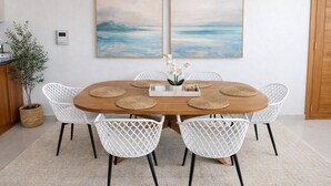 Een ronde houten tafel met witte geweven tafelmatten en witte stoelen met zwarte poten. Daarboven hangen twee ingelijste schilderijen, links staat een potplant en aan de muur een witte thermostaat.