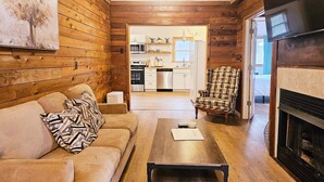 Un accogliente soggiorno con soffitto in legno, camino, divano, poltrona a quadri e una cucina sullo sfondo.