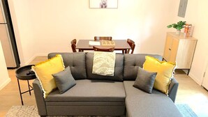 A modern living room with a grey sectional sofa, yellow and grey cushions, a dining table with chairs, a small side table, and a cabinet with a plant. - Central MSU Explorer`s Nest 2BR (Starkville)