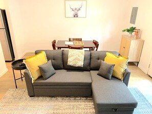 A modern living room with a grey sectional sofa, yellow and grey cushions, a dining table with chairs, a small side table, and a cabinet with a plant. - Central MSU Explorer`s Nest 2BR (Starkville)