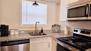 A kitchen with a stainless steel dishwasher, a stove, a microwave, and a sink. There are cabinets, a window with blinds, and a countertop with kitchen utensils. - Cozy 2-Bedroom Haven: Sleeps 5! (Starkville)