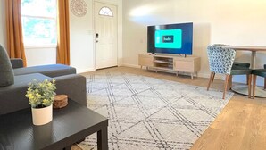 Un salon comprenant un canapé, une table basse avec une plante, un téléviseur posé sur un meuble, une table à manger avec des chaises et un tapis à motifs.