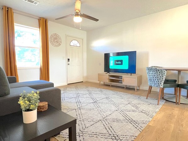 A living room with a sofa, a coffee table with a plant, a TV on a stand, a dining table with chairs, and a patterned rug.