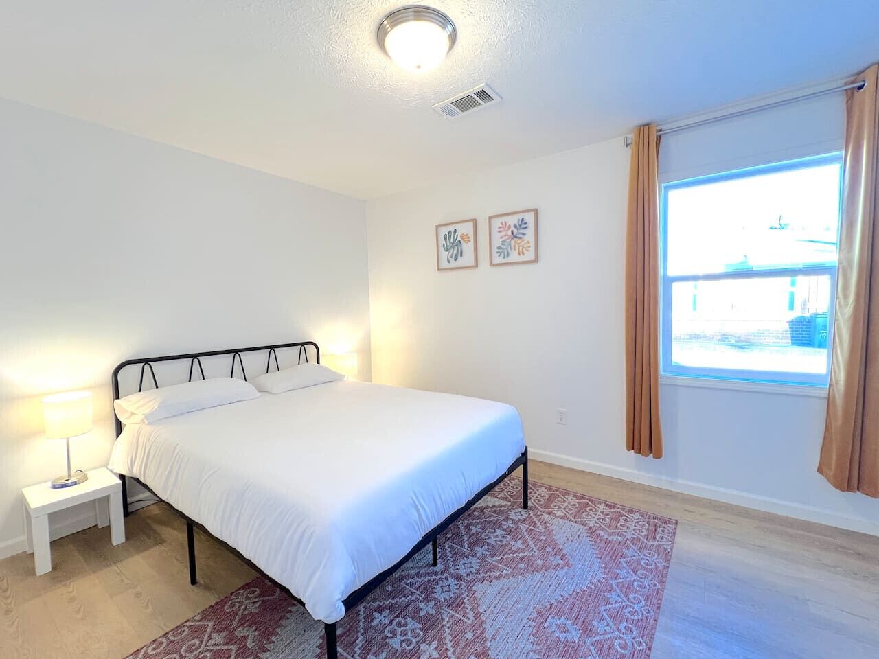 Une chambre à coucher avec un lit, une table de chevet avec une lampe, un tapis à motifs, une fenêtre avec des rideaux et deux œuvres d’art murales.