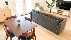 A living room with a wooden dining table, a grey sofa, a green armchair, a TV on a wooden stand, and a potted plant. - MSU Sweet Spot: Cozy 2BR w/Patio (Starkville)