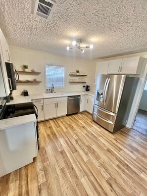 A modern kitchen with white cabinets, a stainless steel refrigerator, and a wooden floor. - Stark Social House | Sleeps 6 | Game Day (Starkville)