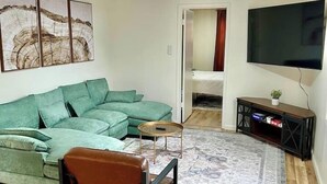 Un salon comprenant un canapé d’angle vert, un fauteuil marron, une table basse, un téléviseur à écran plat et un ventilateur de plafond.