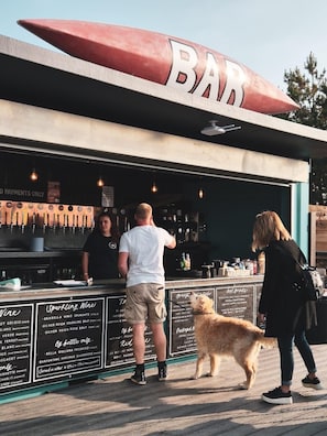 A bar with a large red canoe-shaped sign, a menu board listing various drinks, and a dog on the sidewalk.