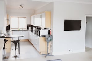 A modern kitchen with white cabinets, a black countertop, and a built-in oven. There are two black bar stools and a standing fan.