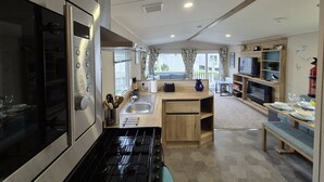 A compact kitchen with a built-in oven, a sink, and a dining area with a TV and seating. - Whitstable Neptune - Alberta Holiday Park (Whitstable)